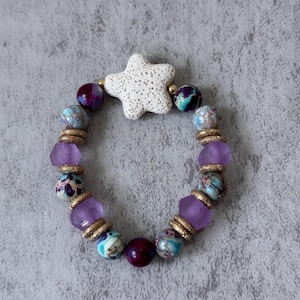 May include: A beaded bracelet featuring a white porous star-shaped bead, interspersed with round purple, teal, and blue patterned beads, translucent light purple beads, and small textured gold-toned spacer beads arranged in a circular pattern.