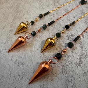 May include: Four pendulums with metallic cone-shaped tips and beaded chains. Two pendulums have gold-colored tips and chains, while the other two have copper-colored tips and chains. The chains feature dark green and black beads.
