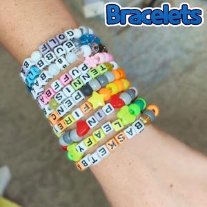 May include: A collection of colorful beaded bracelets spelling out words like "GOLF", "BUBB", "TENNIS", and "LEAFY". The bracelets are made with white letter beads and various colored beads. The image also includes the text "BFDI Themed Bracelets".