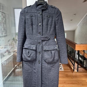 Puede incluir: Un abrigo gris y negro con estampado de pata de gallo, cuello alto, botones y dos bolsillos delanteros. El abrigo tiene una cintura entallada y mangas largas. El abrigo está colgado de un gancho blanco.