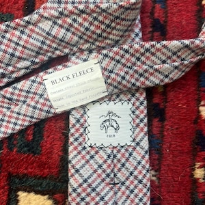 May include: A houndstooth patterned tie in black, red, and white. The label reads "BLACK FLEECE" and "1818". The tie is made of imported fabric and hand-finished in the USA.