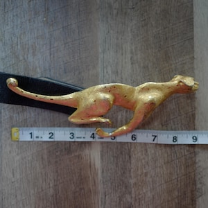 May include: A black leather belt with a gold-colored cheetah buckle. The cheetah buckle measures approximately 4 inches in length. The belt is laid on a wooden surface.