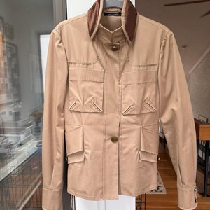 May include: A tan jacket with a brown velvet collar, four front pockets, and button closures. The long-sleeved jacket is hanging on a hanger against a white door.