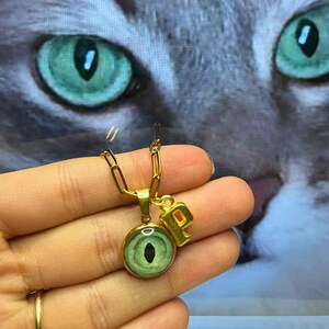 May include: A gold chain necklace with a green eye charm and a gold letter "P" charm. The necklace is being held by a hand.