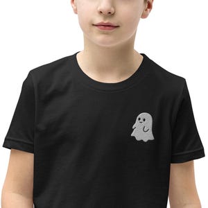 May include: A black short-sleeved t-shirt featuring a small, embroidered light gray ghost graphic on the left chest. The ghost has a simple, rounded shape with two small black eyes and a subtle smile, positioned against the solid black fabric.