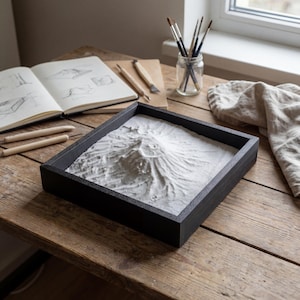 Op de afbeelding: Een zwarte vierkante zandbak met een bergachtig ontwerp in wit zand staat op een houten tafel. Kunstbenodigdheden, waaronder potloden, een schetsboek en penselen in een glazen pot, omringen de bak.