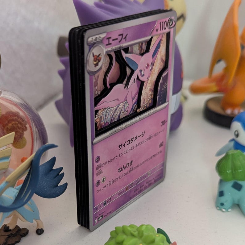 May include: A stack of Pokemon trading cards featuring a pink Espeon character. The cards have Japanese text and are displayed on a white surface with other figurines in the background. The Espeon card has a purple border.