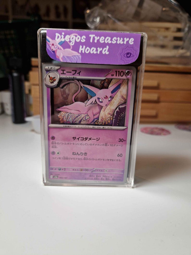 May include: A Pokemon trading card featuring a pink Espeon character, displayed in a clear protective case. The card has Japanese text and a purple border. The case has a purple label that reads "Diegos Treasure Hoard".