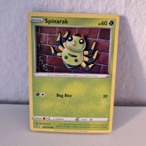 Könnte beinhalten: Eine Spinarak Pokémon-Sammelkarte mit grünem Rand. Die Karte zeigt eine Cartoon-Spinne mit gelbem Körper, blauen Markierungen und schwarzen Beinen. Die Karte trägt den Text "Spinarak", "HP60" und "Bug Bite 30".