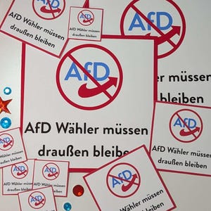 Könnte beinhalten: Eine Ansammlung weißer und roter Schilder mit dem Text "AfD Wähler müssen draußen bleiben." Die Schilder zeigen einen roten Kreis mit einem Strich durch die Buchstaben "AfD" und einen roten Nike-Haken. Einige Schilder sind kleine Quadrate, andere größer.