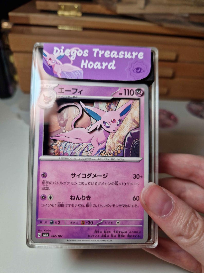 May include: A graded Pok&eacute;mon card featuring Espeon, with Japanese text and a purple and pink color scheme. The card is encased in a clear protective case with a purple label that reads "Diegos Treasure Hoard".