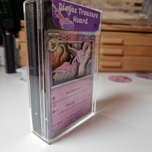 May include: A stack of trading cards in a clear plastic case. The top card features a pink and purple illustration of a creature. The case has a purple label that reads "Diegos Treasure Hoard".