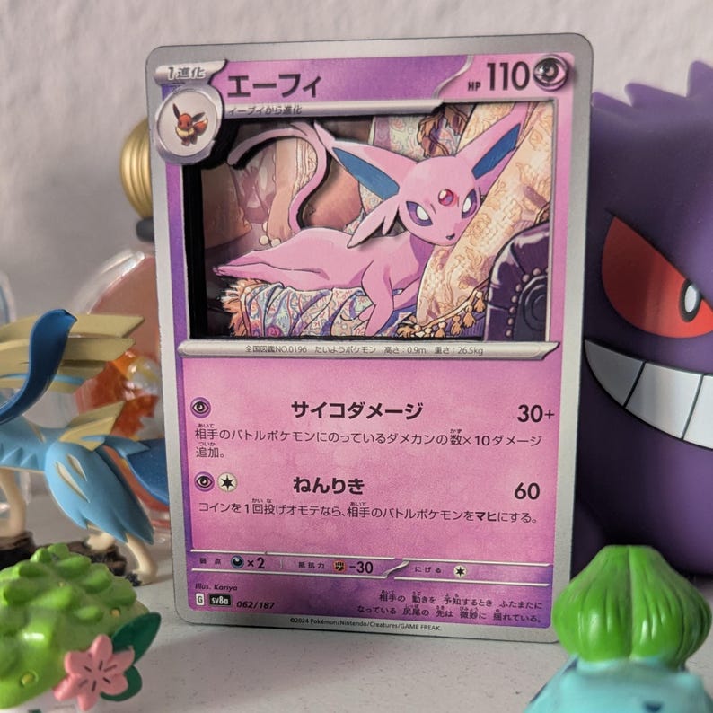 May include: A holographic Pok&eacute;mon trading card featuring a pink Espeon illustration. The card has Japanese text and a HP of 110. The card is surrounded by other Pok&eacute;mon figures.