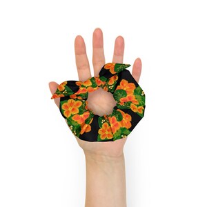 May include: A black scrunchie with a floral pattern of orange flowers and green leaves. The scrunchie is held in a hand against a white background. The fabric is a dark black with a repeating pattern of orange flowers.