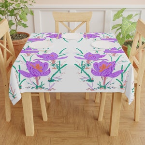 May include: A white tablecloth featuring a repeating pattern of purple crocus flowers with orange centers and green stems. The tablecloth is set on a wooden table with four white plates.