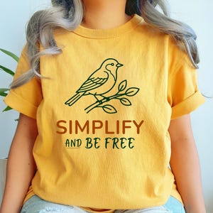 May include: A mustard yellow t-shirt featuring a dark green line art illustration of a bird perched on a branch. Below the graphic, the text reads SIMPLIFY in brown and AND BE FREE in dark green, printed in a clean, sans-serif font style.