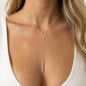 May include: A silver lariat necklace featuring a delicate chain that passes through a small circular ring, ending in a slender vertical bar pendant. The necklace rests against the chest, showcasing a polished metallic finish and a simple design.