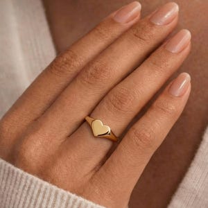 Op de afbeelding: Een goudkleurige ring met een hartvormig ontwerp. De ring wordt om een vinger gedragen, waarbij het hart het centrale punt is. De ring lijkt van gepolijst metaal te zijn, dat licht reflecteert. Een eenvoudig, elegant sieraad.