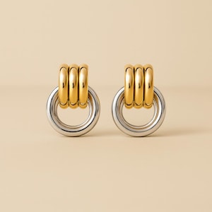 May include: A pair of gold and silver earrings. Each earring features a gold-colored, stacked ring design above a silver-colored, circular hoop. The earrings are displayed on a light beige background.