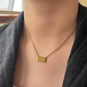 Puede incluir: Un collar de cadena dorada con un colgante rectangular que dice "333" en una fuente estilizada.