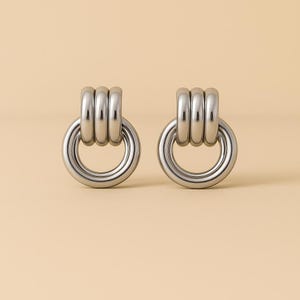 Peut inclure: Une paire de boucles d'oreilles argentées. Chaque boucle d'oreille est composée d'une grande boucle circulaire reliée à un petit motif à trois anneaux superposés. Les boucles d'oreilles ont une finition métallique polie, sur fond neutre.