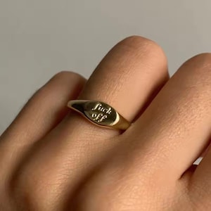 May include: A gold-colored signet ring with the words "fuck off" engraved on the oval face. The ring is worn on a finger, showcasing its polished finish and bold statement.
