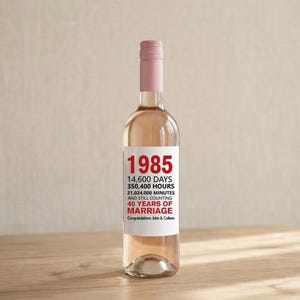 Op de afbeelding: Een heldere wijnfles met een roze dop en een etiket met het jaar "1985" in het rood. Het etiket bevat ook tekst over 40 jaar huwelijk. De fles is gevuld met een lichtroze vloeistof en staat op een houten oppervlak.