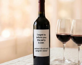 Op de afbeelding: Een donkere glazen wijnfles met een wit rechthoekig etiket met de zwarte tekst: I regret to inform you the party is over. welcome to the real world congrats name! De fles staat opgesteld tussen twee heldere wijnglazen.