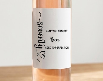 May include: A clear glass wine bottle with a pinkish-orange liquid inside. The label reads "seventy" in cursive, with "Happy 70th Birthday Rocco Aged to Perfection" in black text. The bottle is on a light brown surface.