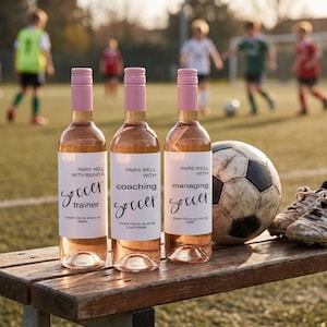 May include: Three glass bottles filled with rosé wine, each featuring a white label with pink screw caps. The labels display the text: PAIRS WELL WITH BEING A soccer trainer, PAIRS WELL WITH coaching soccer, and PAIRS WELL WITH managing soccer.