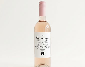 Puede incluir: Una botella de vidrio transparente llena de un líquido rosa claro, con un tapón rosa pálido. La etiqueta rectangular blanca presenta el texto negro New beginnings, memories, adventures, Congratulations, Joe & Kim y un icono de casa negro.