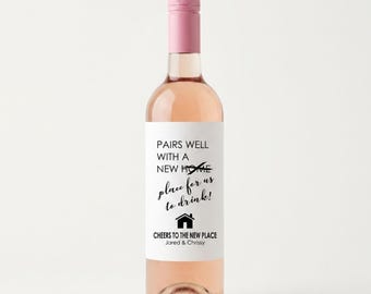 Puede incluir: Una botella de vino de cristal llena de un líquido rosa claro, con un tapón rosa. La etiqueta blanca presenta texto negro que dice: PAIRS WELL WITH A NEW HOME, place for us to drink!, un icono de casa, CHEERS TO THE NEW PLACE y Jared & Chrissy.