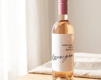 May include: A clear glass wine bottle with a pink cap and a white label. The label reads "PAIRS WELL WITH BEING A Groomsman" in black script with a small heart. The bottle is filled with a light pink liquid.