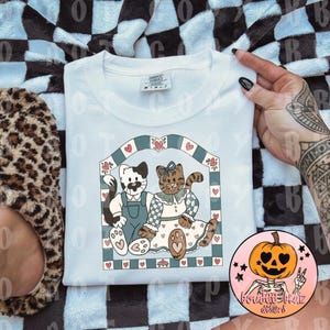 Puede incluir: Sudadera blanca con una ilustración caprichosa de dos gatos dentro de un marco decorativo. Un gato lleva un peto, el otro un vestido. El diseño incluye corazones y un borde azul y blanco. Una pegatina con temática de calabaza es visible.
