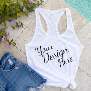 May include: White tank top with the text "Your Design Here" printed on the front. The tank top is laying on a tiled surface with a pair of blue denim shorts and green plants.