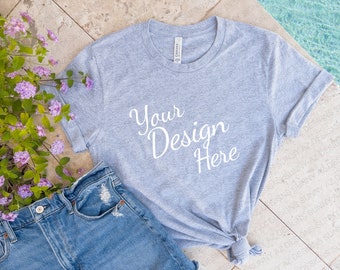 May include: A gray t-shirt with the text "Your Design Here" printed on the front. The t-shirt is lying on a tiled surface with a pair of blue jeans and a bouquet of purple flowers.