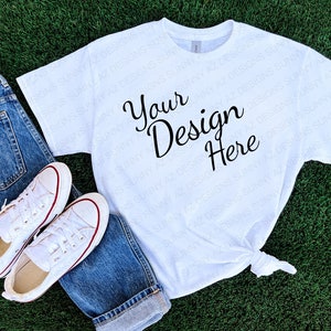 May include: White t-shirt with the text "Your Design Here" printed on the front. The t-shirt is lying on a green grassy surface with a pair of white sneakers and blue jeans.