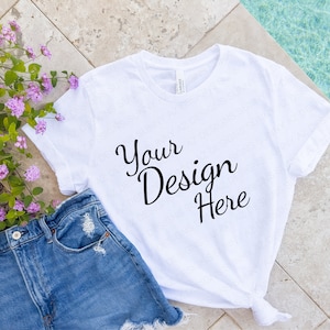 Op de afbeelding: Wit T-shirt met de tekst "Your Design Here" op een betegelde ondergrond met een blauwe denim short en een bos paarse bloemen.