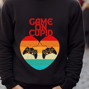 May include: Black sweatshirt with a red heart graphic featuring two video game controllers connected by a cord. The text "GAME ON CUPID" is above the heart.