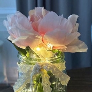 May include: A mason jar with a pink flower arrangement and white lace ribbon. The jar is filled with fairy lights.