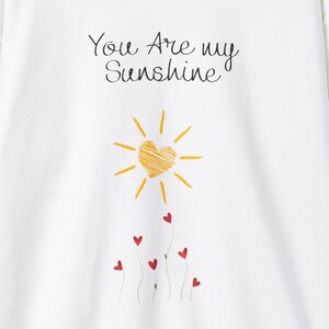 May include: White t-shirt with the text "You Are my Sunshine" and a yellow sun with a heart in the center.  Red hearts are drawn on stems below the sun.