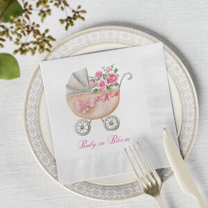 Puede incluir: Una servilleta de papel blanca con una ilustración de acuarela de un cochecito de bebé con rosas rosas y un lazo rosa. El texto "Baby in Bloom" está impreso debajo del cochecito.