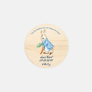 May include: A round wooden plaque with a watercolor illustration of Peter Rabbit holding a carrot. The plaque has the text "Even the Smallest One can change the World" and the baby's name, birthdate, and weight: "Levi Carl 04.24.2024 6 lbs 7 oz".