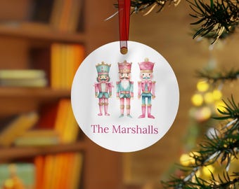 May include: A round white Christmas ornament with three watercolor nutcracker figures. The nutcrackers are wearing pink, blue, and green uniforms. The ornament has the text "The Marshalls" printed on the bottom.