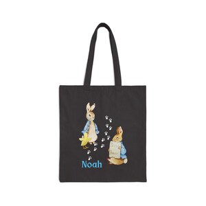 May include: Black canvas tote bag with a Peter Rabbit design featuring two rabbits, one holding a sign that says "Peter Rabbit", and the name "Noah" printed in blue.