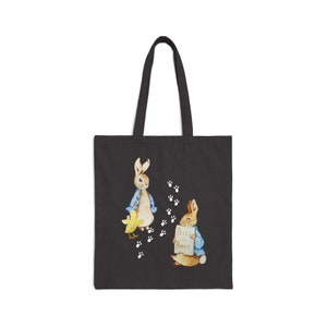 May include: A black canvas tote bag with a white print of two rabbits and a chick. The rabbits are wearing blue shirts and one is holding a sign that says "Peter Rabbit".