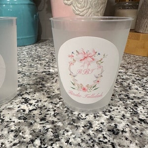 Pode incluir: Dois copos de plástico transparentes com um adesivo branco em cada um. O adesivo tem um laço rosa, flores e o texto "B.B.T. Birdie Tallulah".