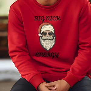 Può includere: Una felpa rossa con una grafica di Babbo Natale con occhiali da sole e barba. Il testo "BIG NICK ENERGY" è stampato sopra e sotto la grafica di Babbo Natale in un carattere nero stilizzato. La felpa ha un girocollo e maniche lunghe.