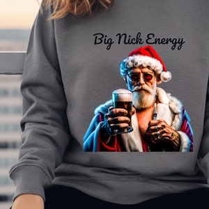 Può includere: Una felpa grigia con la scritta "Big Nick Energy" stampata sul davanti. Il design presenta un'immagine di cartone animato di Babbo Natale con occhiali da sole, che tiene una birra e una sigaretta.