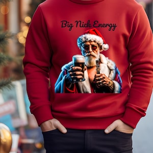 May include: A red sweatshirt featuring the phrase "Big Nick Energy" above an image of Santa Claus in sunglasses, holding a pint of beer and a cigar. The Santa image has a tattoo. The person is wearing black trousers and has their hands in their pockets.